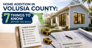 Architectural blueprints, a tape measure, and a building permit resting on a desk in front of a modern home addition in Volusia County, Florida.