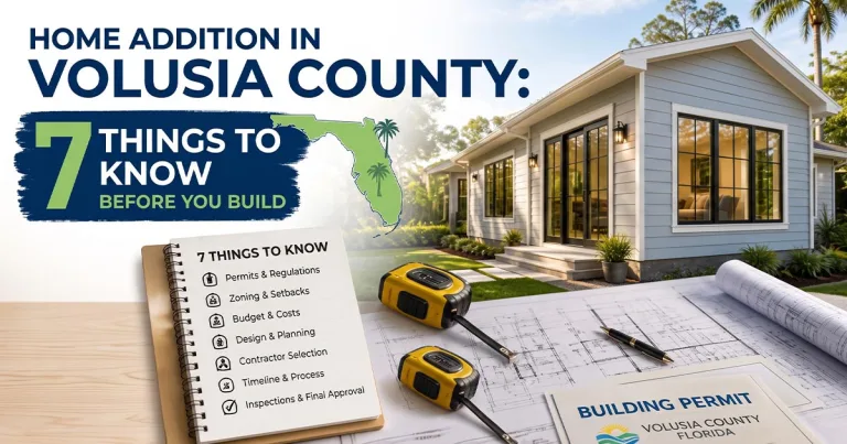 Architectural blueprints, a tape measure, and a building permit resting on a desk in front of a modern home addition in Volusia County, Florida.
